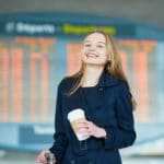 Eine junge Frau steht in einer großen Flughafen Abfertigungshalle und strahlt deutliche Zufriedenheit aus. Mit dem Programm der unibee Institute hat sie deutlich das Selbstbewusstsein gestärkt, somit hat sie jetzt keine Anst mehr, allein zu verreisen. Sie hat lange blonde Haare, einen Becher Kaffee in der Hand und trägt einen dunkelblauen Mantel. Im Hintergrund ist die Anzeigetafel der Abflüge in orangener Schrift zu sehen. Eine junge Frau steht in einer großen Flughafen Abfertigungshalle und strahlt deutliche Zufriedenheit aus. Mit dem Programm der unibee Institute hat sie deutlich das Selbstbewusstsein gestärkt, somit hat sie jetzt keine Anst mehr, allein zu verreisen. Sie hat lange blonde Haare, einen Becher Kaffee in der Hand und trägt einen dunkelblauen Mantel. Im Hintergrund ist die Anzeigetafel der Abflüge in orangener Schrift zu sehen.