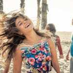 Eine junge Frau in einem Sommerkleid mit bunten Blumen darauf, steht an einem Strand mit Palmen und lacht sehr glücklich. Sie macht einen sehr selbstbewusstsen Eindruck. Sie hat lange braune Haare, welche im Wind wehen. Im Hintergrund sind 3 weitere Personen zu sehen, welche locker tanzen. Eine junge Frau in einem Sommerkleid mit bunten Blumen darauf, steht an einem Strand mit Palmen und lacht sehr glücklich. Sie macht einen sehr selbstbewusstsen Eindruck. Sie hat lange braune Haare, welche im Wind wehen. Im Hintergrund sind 3 weitere Personen zu sehen, welche locker tanzen.