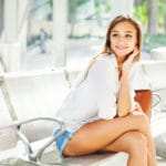 Eine junge Frau sitzt in einem Wartesaal und lächelt zufrieden. Sie strahlt vollstes Selbstbewusstsein aus. Sie hat lange blonde Haare und trägt eine weisse Bluse mit einer kurzen Jeanshose. Mit ihrem linken Arm fasst sie sich hinter das Ohr und stützt den Kopf leicht ab. Eine junge Frau sitzt in einem Wartesaal und lächelt zufrieden. Sie strahlt vollstes Selbstbewusstsein aus. Sie hat lange blonde Haare und trägt eine weisse Bluse mit einer kurzen Jeanshose. Mit ihrem linken Arm fasst sie sich hinter das Ohr und stützt den Kopf leicht ab.