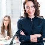 Eine Geschäftsfrau steht mit verschränkten Armen vor einem Schreibtisch. Damit zeigt sie, dass sie mit dem Programm der unibee Institute, ein gutes Selbstbewusstsein gewinnen konnte. Ihr lächeln zeigt eine ausgeglichene und sichere Lebensbalance. Sie ist bekleidet mit einer dunkelblauen Jacke und trägt lockige braune Haare, welche Schulterlang sind. Im Hintergrund sind 3 andere Personen verschwommen zu sehen, weil der Kamerafokus ausschließlich auf die Frau bezogen ist. Eine Geschäftsfrau steht mit verschränkten Armen vor einem Schreibtisch. Damit zeigt sie, dass sie mit dem Programm der unibee Institute, ein gutes Selbstbewusstsein gewinnen konnte. Ihr lächeln zeigt eine ausgeglichene und sichere Lebensbalance. Sie ist bekleidet mit einer dunkelblauen Jacke und trägt lockige braune Haare, welche Schulterlang sind. Im Hintergrund sind 3 andere Personen verschwommen zu sehen, weil der Kamerafokus ausschließlich auf die Frau bezogen ist.