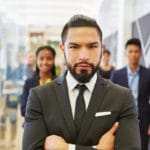 Ein Mann steht in einem Großraumbüro und schaut mit verschränkten Armen direkt in die Kamera. Er strahlt großartiges Selbstbewusstsein aus welches er mit dem Programm der unibee Institute gewonnen hat. Er ist bekleidet mit einem grauen Anzug und grauer Kravatte auf weissen Hemd. Er hat kurze schwarze Haare und trägt einen Vollbart. Im Hintergrund 4 Menschen verschwommen zu sehen, weil der Kamerafokus nur auf den Geschäftsmann gerichtet ist. Ein Mann steht in einem Großraumbüro und schaut mit verschränkten Armen direkt in die Kamera. Er strahlt großartiges Selbstbewusstsein aus welches er mit dem Programm der unibee Institute gewonnen hat. Er ist bekleidet mit einem grauen Anzug und grauer Kravatte auf weissen Hemd. Er hat kurze schwarze Haare und trägt einen Vollbart. Im Hintergrund 4 Menschen verschwommen zu sehen, weil der Kamerafokus nur auf den Geschäftsmann gerichtet ist.