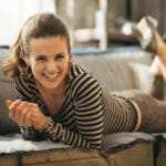 Eine Frau liegt auf einem Sofa auf dem Bauch. Sie lächelt sehr freundlich und zeigt mit der Gesichtsmimik, dass das Programm der unibee Institute zum Selbstbewusstsein stärken, sehr gute Erfolge gebracht hat. Sie ist bekleidet mit einem grau/beige getreiften Pullover, einer grauen Jeanshose und silbergrauen Damenschuhe mit hohen Absätzen. Um den Arm hat sie viele Goldketten als Modeschmuck. Ihre Haare sind dunkelbraun, welche zu einem Zopf nach hinten zusammengebunden sind. Eine Frau liegt auf einem Sofa auf dem Bauch. Sie lächelt sehr freundlich und zeigt mit der Gesichtsmimik, dass das Programm der unibee Institute zum Selbstbewusstsein stärken, sehr gute Erfolge gebracht hat. Sie ist bekleidet mit einem grau/beige getreiften Pullover, einer grauen Jeanshose und silbergrauen Damenschuhe mit hohen Absätzen. Um den Arm hat sie viele Goldketten als Modeschmuck. Ihre Haare sind dunkelbraun, welche zu einem Zopf nach hinten zusammengebunden sind.