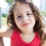 Auf diesem Bild ist ein kleines Mädchen im Alter von ungefähr 6 Jahren zu sehen. Es hat lange braune, gelockte Haare und ist mit einem roten Ärmellosen Shirt bekleidet und es ist ein Sommertag. Das Kind strahlt mit der Gesichtsmimik und Körperhatung ein gutes Selbstbewusstsein aus. Auf diesem Bild ist ein kleines Mädchen im Alter von ungefähr 6 Jahren zu sehen. Es hat lange braune, gelockte Haare und ist mit einem roten Ärmellosen Shirt bekleidet und es ist ein Sommertag. Das Kind strahlt mit der Gesichtsmimik und Körperhatung ein gutes Selbstbewusstsein aus.