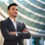 Ein junger Man mit kurzen schwarzen Haaren steht mit verschränkten Armen und erhobenem Kopf vor einem großen Bürogebäude. Er strahlt mit seiner Mimik ein starkes Selbstbewusstsein aus. Er ist bekleidet mit einem grauen Anzug und weissem Hemd. Ein junger Man mit kurzen schwarzen Haaren steht mit verschränkten Armen und erhobenem Kopf vor einem großen Bürogebäude. Er strahlt mit seiner Mimik ein starkes Selbstbewusstsein aus. Er ist bekleidet mit einem grauen Anzug und weissem Hemd.