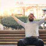 Ein junger Mann mit Vollbart und kurzen braunen Haaren, sitzt auf einer Bank an einer Strasse vor einem alten großen Gebäude. Er hebt beide Arme nach oben und ballt seine Hände zu einer Faust und lächelt dabei sehr glücklich. Er zeigt mit seiner Gestik und Gesichtsmimik, dass er mit dem Programm der unibee Institute, sein Selbstbewusstsein ausserordentlich gut aufbauen konnte. Er ist bekleidet mit einem blauen Hemd und einer schwarzen Jeanshose. Es ist ein sonniger Tag. Ein junger Mann mit Vollbart und kurzen braunen Haaren, sitzt auf einer Bank an einer Strasse vor einem alten großen Gebäude. Er hebt beide Arme nach oben und ballt seine Hände zu einer Faust und lächelt dabei sehr glücklich. Er zeigt mit seiner Gestik und Gesichtsmimik, dass er mit dem Programm der unibee Institute, sein Selbstbewusstsein ausserordentlich gut aufbauen konnte. Er ist bekleidet mit einem blauen Hemd und einer schwarzen Jeanshose. Es ist ein sonniger Tag.