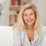 Eine ältere Frau ungefähr 50 Jahre mit langen blonden Haaren lächelt zufrieden und beugt sich leicht nach vorn zur Kamera hin. Mit ihrer Mimik strahlt sie gute Balance und gutes, stabiles Selbstbewusstsein aus. Sie sitzt auf einem weissen Sofa welches in einem Wohnzimmer steht. Im Hintergrund ist ein Bücherregal mit orangenen Büchern zu sehen und links daneben steht eine große grüne Zimmerpflanze. Eine ältere Frau ungefähr 50 Jahre mit langen blonden Haaren lächelt zufrieden und beugt sich leicht nach vorn zur Kamera hin. Mit ihrer Mimik strahlt sie gute Balance und gutes, stabiles Selbstbewusstsein aus. Sie sitzt auf einem weissen Sofa welches in einem Wohnzimmer steht. Im Hintergrund ist ein Bücherregal mit orangenen Büchern zu sehen und links daneben steht eine große grüne Zimmerpflanze.