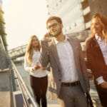 Ein Mann im grauen Anzug und weissen Hemd geht auf der Strasse entlang und telefoniert dabei lächelnd mit seinem Handy. Er hat kurze braune Haare und einen Vollbart. Neben Ihm läuft eine Frau mit sehr langen blonden Haaren, die ihn freundlich anlächelt. Der Mann zeigt deutlich sein gewonnens Selbstbewusstsein und seine Mimik zeigt deutlich, dass er Lust am arbeiten hat. Im Hintergrund ist ein graues Fabrikgebäude zu sehen. Es ist ein trüber Wolkenbehangener Tag. Die Frau an der Seite des Mannes ist bekleidet mit einem schwarzen Anzug und weissen Hemd mit Stehkragen. Ein Mann im grauen Anzug und weissen Hemd geht auf der Strasse entlang und telefoniert dabei lächelnd mit seinem Handy. Er hat kurze braune Haare und einen Vollbart. Neben Ihm läuft eine Frau mit sehr langen blonden Haaren, die ihn freundlich anlächelt. Der Mann zeigt deutlich sein gewonnens Selbstbewusstsein und seine Mimik zeigt deutlich, dass er Lust am arbeiten hat. Im Hintergrund ist ein graues Fabrikgebäude zu sehen. Es ist ein trüber Wolkenbehangener Tag. Die Frau an der Seite des Mannes ist bekleidet mit einem schwarzen Anzug und weissen Hemd mit Stehkragen.