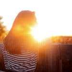 Eine Frau steht auf einer Terasse und sieht einen leuchtend hellen Sonnenuntergang zu. Auf dem Foto ist die Frau von hinten zu sehen und sie ist bekleidet mit einem weiss/blau getreiften Pullover. Sie hat lange braune Haare. Sie versucht durch den Blick in den Sonnenuntergang um ihre kreisenden Gedanken loszulassen um ihr Selbstbewusstsein zu spüren und zu stärken. Eine Frau steht auf einer Terasse und sieht einen leuchtend hellen Sonnenuntergang zu. Auf dem Foto ist die Frau von hinten zu sehen und sie ist bekleidet mit einem weiss/blau getreiften Pullover. Sie hat lange braune Haare. Sie versucht durch den Blick in den Sonnenuntergang um ihre kreisenden Gedanken loszulassen um ihr Selbstbewusstsein zu spüren und zu stärken.