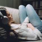 Eine junge Frau liegt entspannt im Sofa und hört eine Hypnose CD zum Selbstbewusstsein stärken von den unibee Instituten. Sie hat lange schwarze Haare und große silberne Kopfhörer auf. Bekleidet ist sie mit einem grauen Pullover und einer hellblauen klassischen Jeanshode. Im Hintergrund ist ein Bücherregal zu sehen, worin sich viele weisse Aktenordner befinden. Eine junge Frau liegt entspannt im Sofa und hört eine Hypnose CD zum Selbstbewusstsein stärken von den unibee Instituten. Sie hat lange schwarze Haare und große silberne Kopfhörer auf. Bekleidet ist sie mit einem grauen Pullover und einer hellblauen klassischen Jeanshode. Im Hintergrund ist ein Bücherregal zu sehen, worin sich viele weisse Aktenordner befinden.