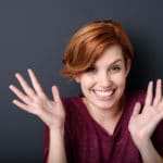 Eine Frau mit rotbraunen kurzen Haaren lächelt und hebt dabei beide Hände hoch. Sie strahlt mit Ihrer Mimik und Gestik vollstes Selbstbewusstsein aus. Sie ist bekleidet mit einem lilafarbenen Pullover. Der Hintergrund ist einfarbig dunkelblau. Eine Frau mit rotbraunen kurzen Haaren lächelt und hebt dabei beide Hände hoch. Sie strahlt mit Ihrer Mimik und Gestik vollstes Selbstbewusstsein aus. Sie ist bekleidet mit einem lilafarbenen Pullover. Der Hintergrund ist einfarbig dunkelblau.